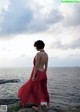 A woman in a red dress standing on a rock by the ocean.